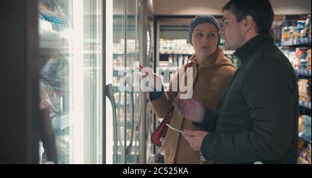 Les jeunes choisissent de la nourriture dans la section frigo du supermarché. Femme prenant deux paquets de fraises congelées, alors que l'homme vérifie la liste de magasin sur le Banque D'Images