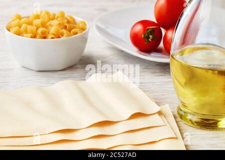 Feuilles de pâtes de lasagne séchées non cuites à l'huile d'olive et aux tomates cerises sur une table de cuisine en bois blanc. Banque D'Images