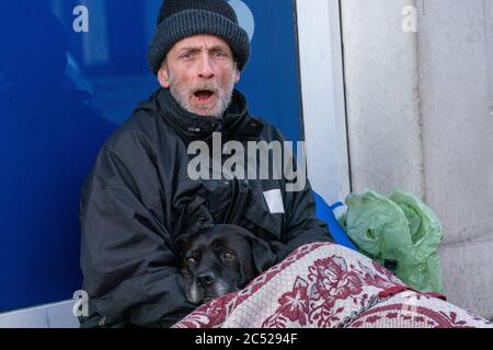 Un homme sans abri âgé, assis avec son dos contre une fenêtre de magasin. Câliner un chien, tout en criant aux gens qui passent. Banque D'Images