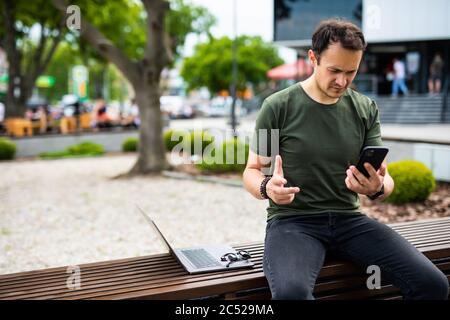 Entrepreneur travaillant la messagerie texte dans un téléphone portable assis sur un banc avec des immeubles de bureau Banque D'Images