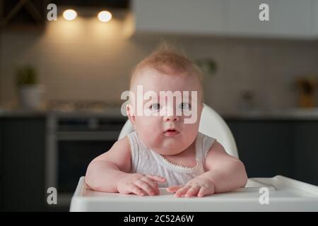 un petit enfant dans un body blanc est assis dans un chaise haute dans la cuisine et attend de manger Banque D'Images