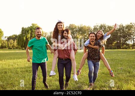 Groupe d'amis marchant dans la campagne avec des gars donnant le pigeyback tour aux filles. Les jeunes s'amusent dans la nature Banque D'Images