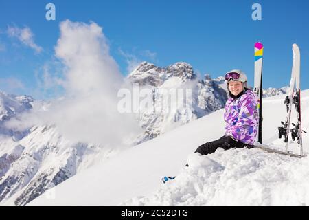 Bonne fille dans la tenue de ski, casque et masque de loin assis dans la neige sur le sommet de montagne regarder la caméra Banque D'Images