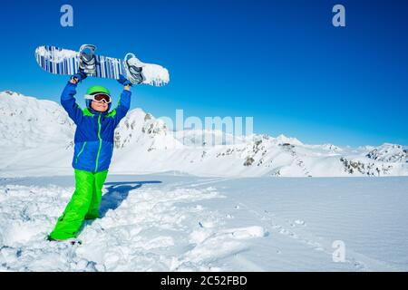 Petit garçon tient le snowboard au-dessus de la tête dans les mains sur la vue de neige du côté dans le masque, casque de ski Banque D'Images