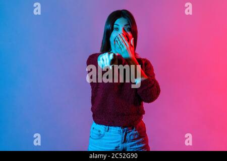 Hé, vous ! Portrait de lumière néon de femme impressionnée choquée pointant le doigt vers l'appareil photo et couvrant la bouche dans étonnement, regardant surpris, surpris par certains Banque D'Images