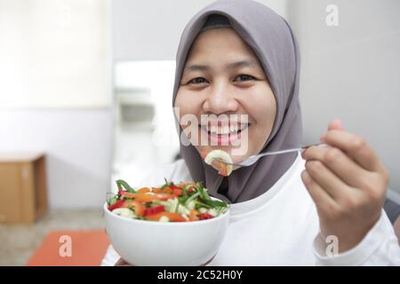 Femme asiatique musulmane portant hijab manger une nourriture saine, salade de légumes verts, après l'exercice à la maison, garder en bonne santé et en forme pendant le nouveau mode de vie normal co Banque D'Images