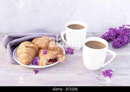 Deux belles tasses de café en porcelaine avec du lait et des croissants décorés de fleurs lilas sur une table en bois blanc. Concept de petit déjeuner parfait. Copier l'espace. Banque D'Images