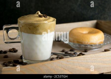 Gros plan du café instantané fouetté sur un verre de lait avec un beignet sur un plateau en bois parsemé de grains de café. Le concept de café Dalgona. Banque D'Images