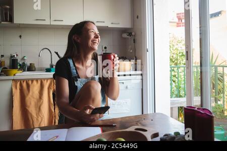 Jeune femme assise sur une table de cuisine en bois avec une atmosphère détendue en vérifiant son smartphone et en écrivant des notes; concept de style de vie naturel, simplicité Banque D'Images