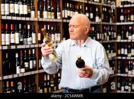 Un homme âgé sérieux choisit entre le vin rouge et le vin blanc dans un magasin de spiritueux Banque D'Images