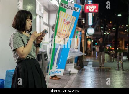 Jeune fille japonaise avec smartphone debout sur le trottoir de la rue Dogenzakue, la nuit, Tokyo, Japon Banque D'Images