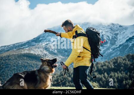 Un jeune homme avec une veste jaune et un sac à dos joue avec le Berger allemand dans les montagnes. Banque D'Images