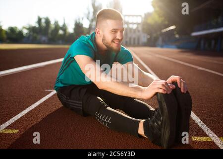 Jeune homme souriant dans le sport porter heureux regardant de côté et s'étirant tout en passant du temps sur le tapis roulant du stade Banque D'Images