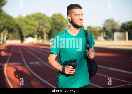 Jeune homme dans des écouteurs sans fil tenant la bouteille de sport à la main regardant avec plaisir sur la piste de course du stade Banque D'Images