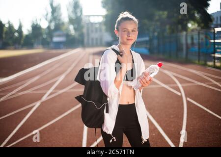 Jolie fille dans le sport porter rêve regardant dans l'appareil photo avec sac à dos sur l'épaule et bouteille d'eau à la main sur la piste de course du stade Banque D'Images