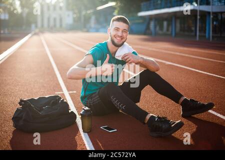 Jeune homme souriant regardant avec joie dans l'appareil photo avec une serviette blanche sur l'épaule montrant le pouce vers le haut tout en passant du temps sur l'hippodrome du stade Banque D'Images