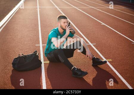 Un jeune homme de sport porte de l'eau potable de façon réfléchie de la bouteille de sport tout en passant du temps sur le tapis roulant du stade Banque D'Images