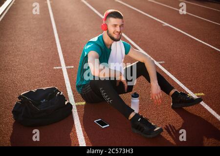 Jeune homme souriant dans des écouteurs rouges regardant avec joie de côté avec une serviette sur l'épaule sur la piste de course du stade Banque D'Images