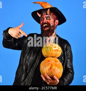 Monster avec décorations d'octobre. Spooky Halloween party concept. Démon avec des cornes et heureux face à jack o lanternes. Homme portant un miroir effrayant et witchers hat détient les citrouilles sur fond bleu Banque D'Images