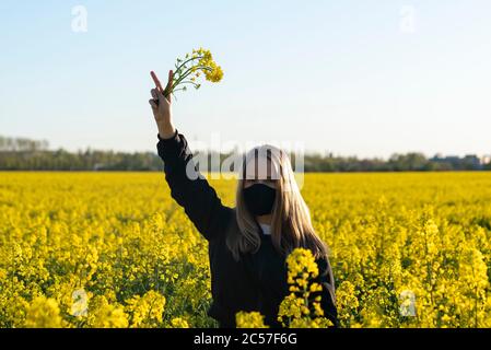 Une jeune fille portant un masque pour se protéger contre les peuplements de corona dans un champ de colza et montre le signe de la victoire Banque D'Images