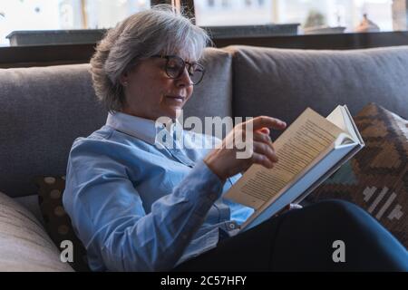 Femme adulte avec des lunettes lisant un livre et tournant la page sur un canapé gris à l'intérieur d'une maison avec la lumière entrant par la fenêtre Banque D'Images