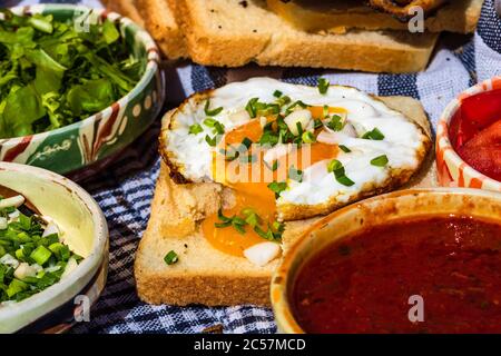Composition rustique avec des petits pains aux saucisses, des œufs frits sur du pain grillé, différents bols avec sauce et des légumes hachés. Banque D'Images