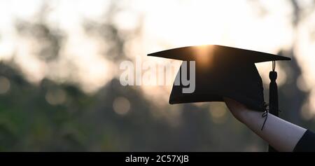 Image rognée de la main d'un étudiant de l'université tenant un chapeau de graduation à la main à l'extérieur avec le coucher du soleil comme arrière-plan. Banque D'Images