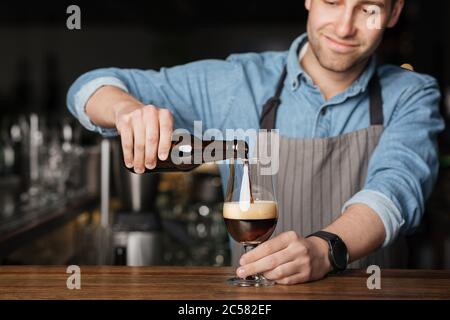 Répétez l'ordre. Barman en tablier et chemise en denim, verse de la bière foncée en verre Banque D'Images