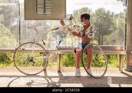 Un jeune homme charmant qui attend à la gare routière. Il a une moto avec un pneu à plat. Il porte un masque et une chemise d'été. Banque D'Images