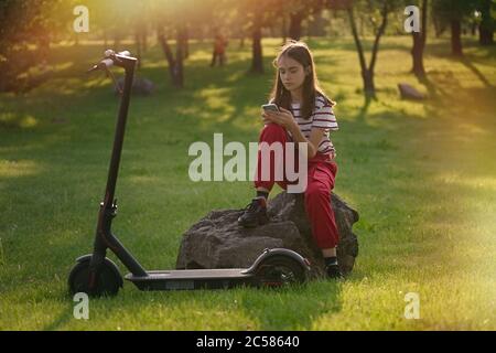 Une jeune fille adorable se détendant après avoir fait du vélo électrique et utilisé son smartphone dans le parc au coucher du soleil Banque D'Images