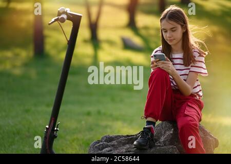 Une jeune fille adorable se détendant après avoir fait du vélo électrique et utilisé son smartphone dans le parc au coucher du soleil Banque D'Images