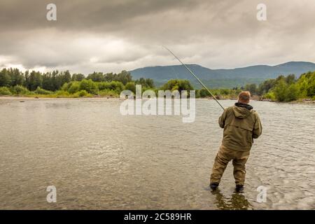 Un pêcheur s'est affronté à un saumon tout en barbotant sur la rivière Kitimat, en Colombie-Britannique, au Canada, avec la canne courbée et la forêt et les montagnes dans l'arrière-gro Banque D'Images