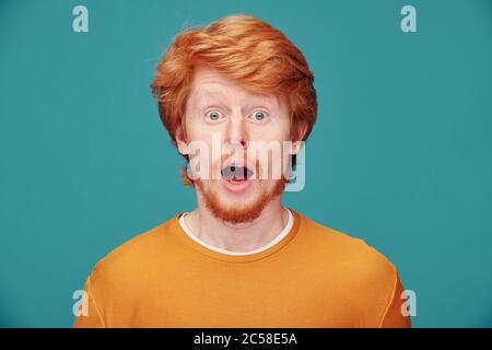 Portrait de jeune homme émotif à barbe rouge surpris par les nouvelles en gardant la bouche ouverte sur fond bleu Banque D'Images