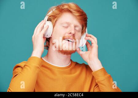 Jeune homme à tête rouge décontracté dans un chandail orange qui garde les yeux fermés tout en écoutant de la musique dans un casque Banque D'Images