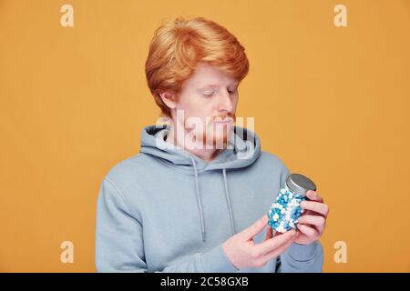 Grave sceptique jeune barbu rouge en lecture à capuchon étiquette sur la bouteille de vitamine sur fond orange Banque D'Images