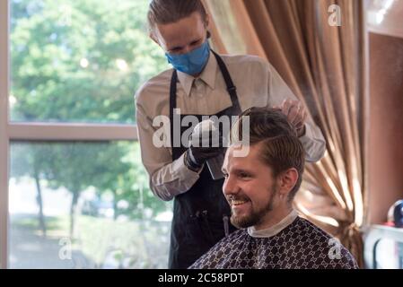 un barbier dans un masque de protection dans un barbershop est en cours de coupe d'un jeune barbu. coupe de cheveux de quarantaine, barbershop travailleur dans un masque de protection rasant un gars Banque D'Images