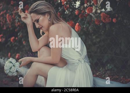 la mariée en robe blanche pleure, tenant une cigarette dans ses mains, tenant une bouteille d'alcool. Sur fond floral de Banque D'Images