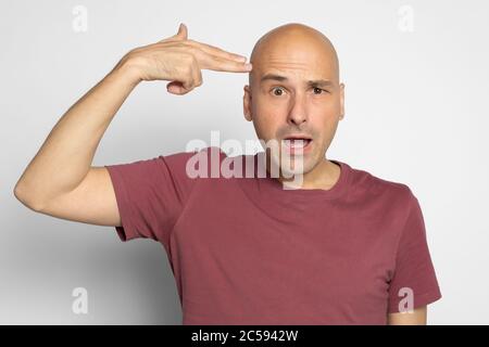 Portrait d'un homme chauve avec un geste de pistolet virtuel sur sa tête. Isolé sur un mur gris Banque D'Images