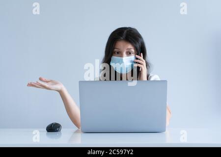 La fille travaille de la maison pendant la quarantaine. Pandémie du virus Corona. Une fille dans un masque chirurgical travaille sur un ordinateur portable et boit du café. Travail à distance. Banque D'Images