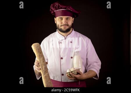 Souriant beau barbu chef homme en uniforme tient la baguette, le biberon de lait, les œufs Banque D'Images