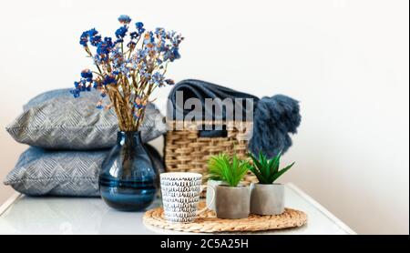 Décoration intérieure chaleureuse : tasse de café, oreiller, écossais, vase avec fleurs, plantes en pots sur un support en osier sur une étagère blanche dans la chambre. Le quaranti Banque D'Images