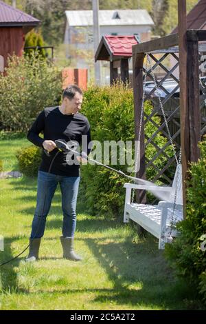 40s homme lave les balançoires blanches dans le jardin de maison de campagne avec un nettoyeur haute pression Banque D'Images