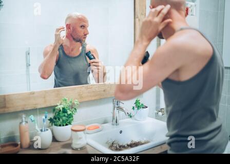 Homme sans cheveux touchant son nouveau style de coupe à tête chauve il a fait à l'aide d'une tondeuse électrique rechargeable regardant dans le miroir de salle de bains. Décision radicale, lif Banque D'Images