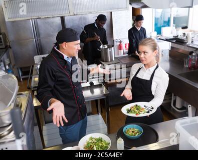 Jeune serveuse debout avec plat cuisiné dans une cuisine de restaurant à parler avec perplexe male chef Banque D'Images