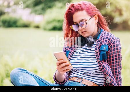 Portrait d'une jeune femme adolescente moderne avec une couleur de coiffure extraordinaire dans une chemise à carreaux avec casque sans fil, envoi de SMS via smartp Banque D'Images