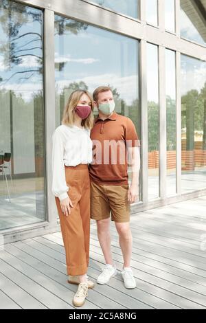 Beau couple en masques en tissu debout contre le cottage et embrassant tout en posant pour la photo Banque D'Images