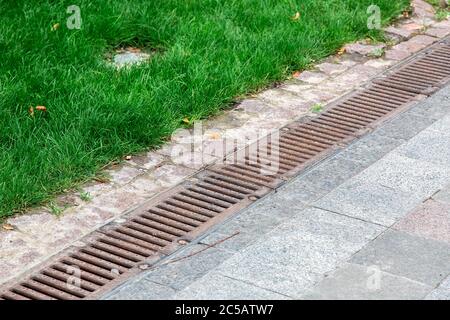 grille de drainage rouillé d'un système de tempête sur un trottoir piétonnier en carreaux de pierre dans un parc près d'une herbe verte. Banque D'Images