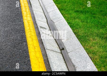 plateau de drainage de bord de route avec eau de pluie sur une route asphaltée avec un marquage jaune et une pelouse verte. Banque D'Images