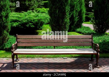 banc en bois brun avec des pieds en fer sur la voie de pied fait de tuiles rouges dans l'arrière-plan espaces verts bien entretenu jardin avec des buissons de thuja et vert Banque D'Images