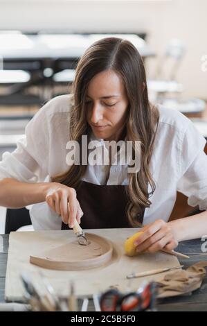 Femme travaillant avec de l'argile avec ses mains et outil de coupe en boucle gros plan dans un atelier de poterie. Banque D'Images